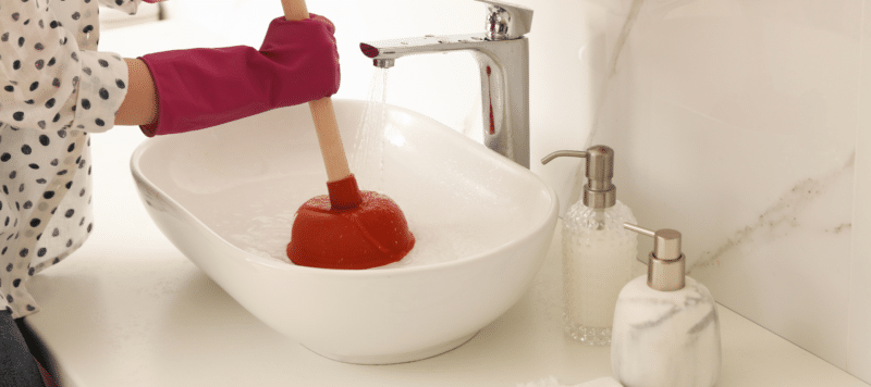 woman with gloved hands using a plunger to unclog a bathroom sink